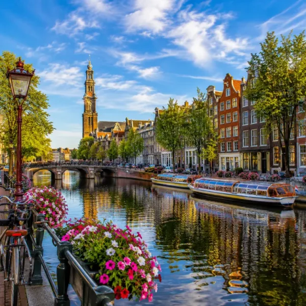 Amsterdam canal with historic houses, tour boats, bicycles, flowers, bridge, and Westerkerk tower in clear daylight.
