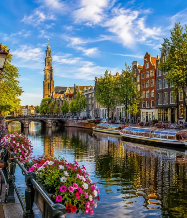 Amsterdam canal with historic houses, tour boats, bicycles, flowers, bridge, and Westerkerk tower in clear daylight.