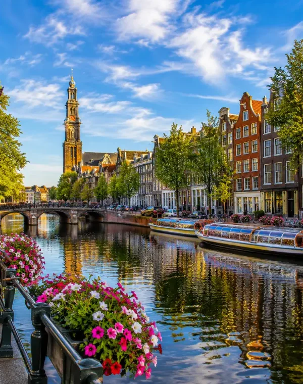 Amsterdam canal with historic houses, tour boats, bicycles, flowers, bridge, and Westerkerk tower in clear daylight.