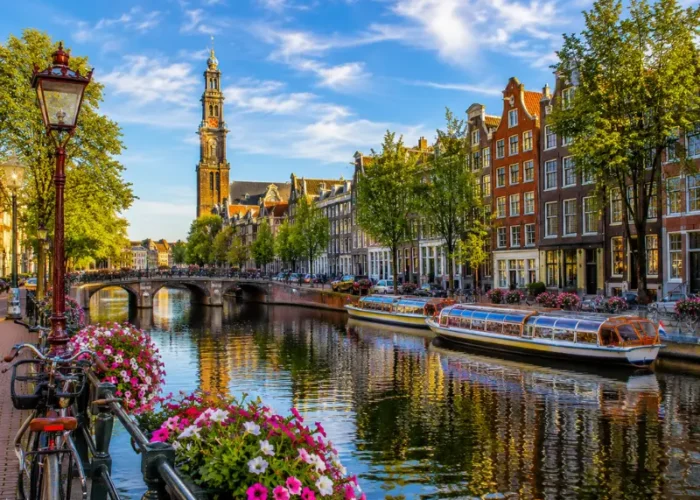 Amsterdam canal with historic houses, tour boats, bicycles, flowers, bridge, and Westerkerk tower in clear daylight.