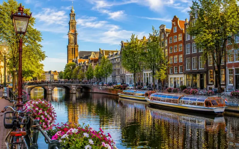 Amsterdam canal with historic houses, tour boats, bicycles, flowers, bridge, and Westerkerk tower in clear daylight.