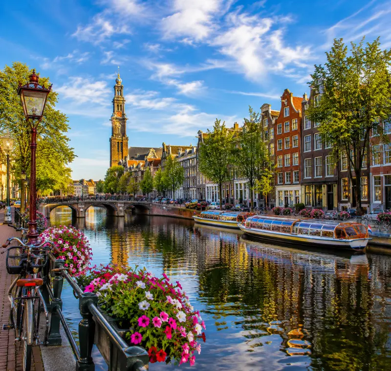 Amsterdam canal with historic houses, tour boats, bicycles, flowers, bridge, and Westerkerk tower in clear daylight.