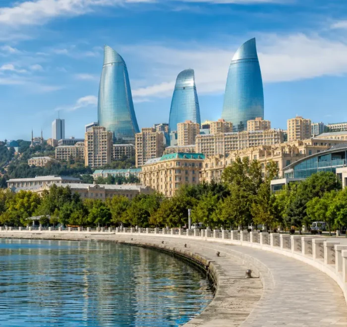 Bright daytime view of Baku waterfront with the iconic Flame Towers, tree-lined promenade, and calm Caspian shoreline.