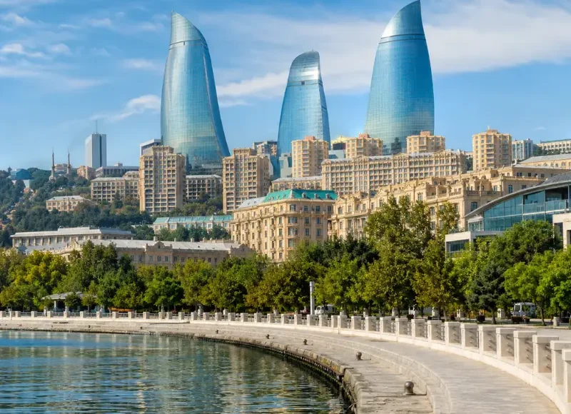 Bright daytime view of Baku waterfront with the iconic Flame Towers, tree-lined promenade, and calm Caspian shoreline.