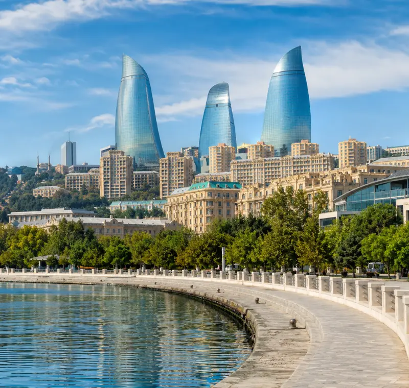 Bright daytime view of Baku waterfront with the iconic Flame Towers, tree-lined promenade, and calm Caspian shoreline.