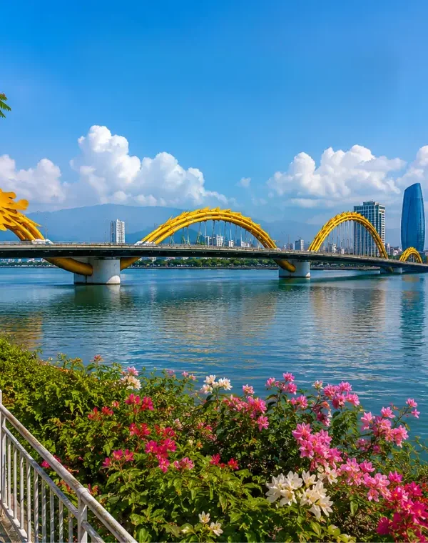 Da Nang, Vietnam skyline with Dragon Bridge, Han River, riverside flowers, boats, and clear blue daytime sky.