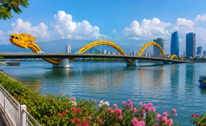 Da Nang, Vietnam skyline with Dragon Bridge, Han River, riverside flowers, boats, and clear blue daytime sky.