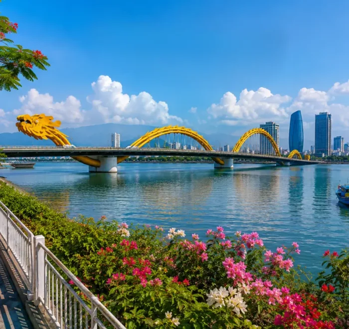 Da Nang, Vietnam skyline with Dragon Bridge, Han River, riverside flowers, boats, and clear blue daytime sky.