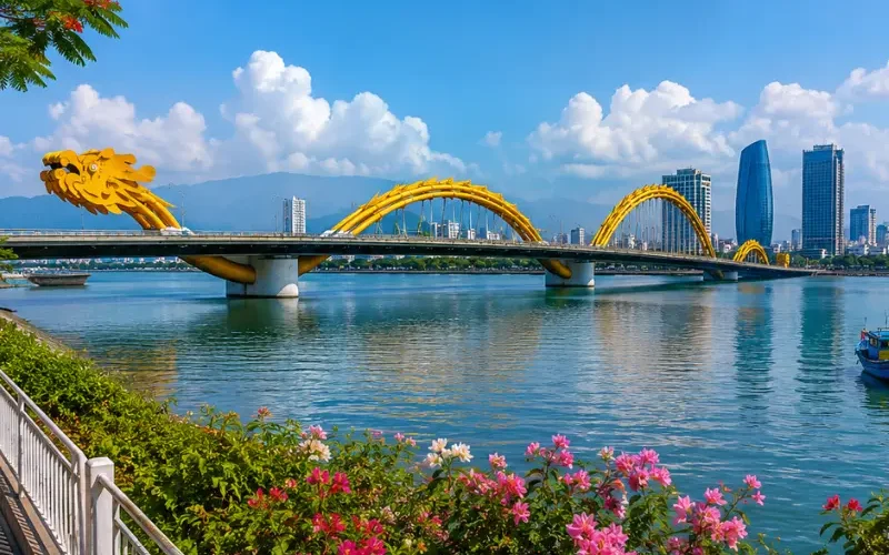 Da Nang, Vietnam skyline with Dragon Bridge, Han River, riverside flowers, boats, and clear blue daytime sky.