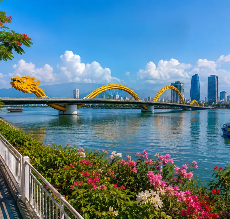 Da Nang, Vietnam skyline with Dragon Bridge, Han River, riverside flowers, boats, and clear blue daytime sky.