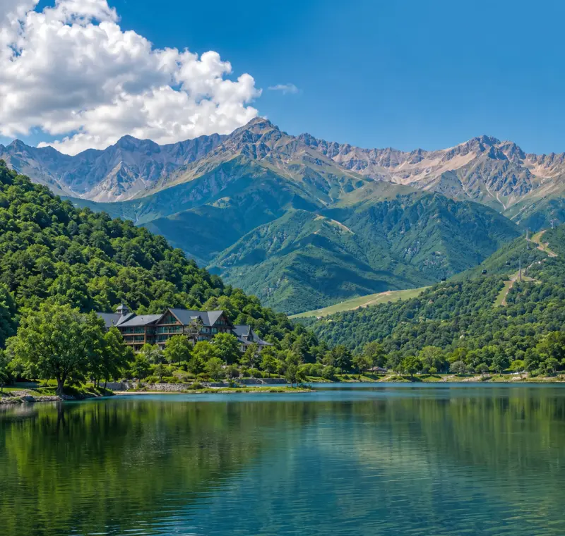 Scenic daytime view of Gabala with a calm lake, lush green hills, mountain resort, and the Caucasus peaks in the background.