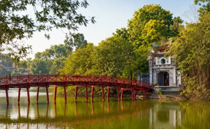 Hoan Kiem Lake in Hanoi with red The Huc Bridge, Ngoc Son Temple, calm water, and lush green trees.