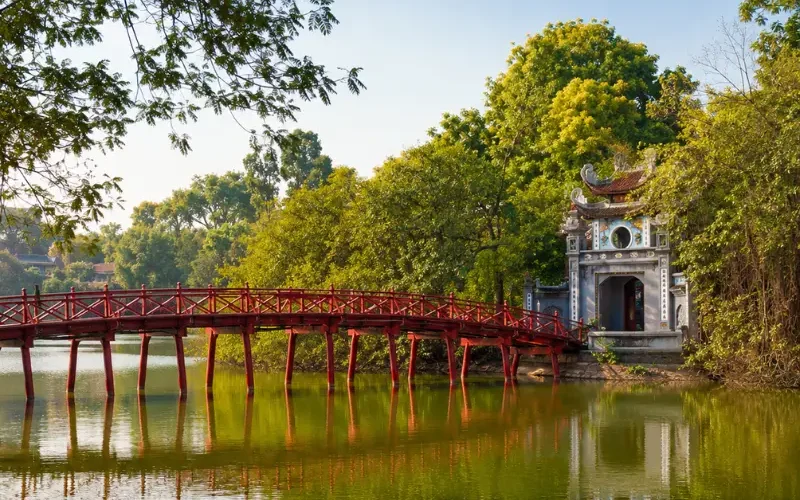 Hoan Kiem Lake in Hanoi with red The Huc Bridge, Ngoc Son Temple, calm water, and lush green trees.