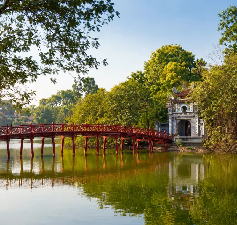 Hoan Kiem Lake in Hanoi with red The Huc Bridge, Ngoc Son Temple, calm water, and lush green trees.