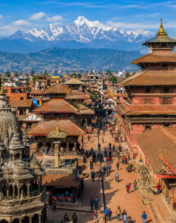Bright daytime view of Kathmandu with traditional temples, historic square, city rooftops, and Himalayan mountains.