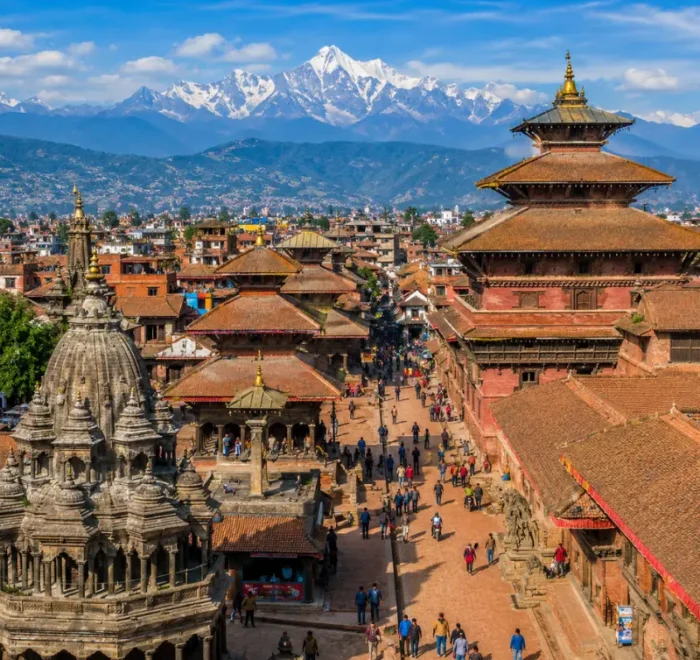 Bright daytime view of Kathmandu with traditional temples, historic square, city rooftops, and Himalayan mountains.