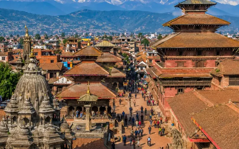 Bright daytime view of Kathmandu with traditional temples, historic square, city rooftops, and Himalayan mountains.