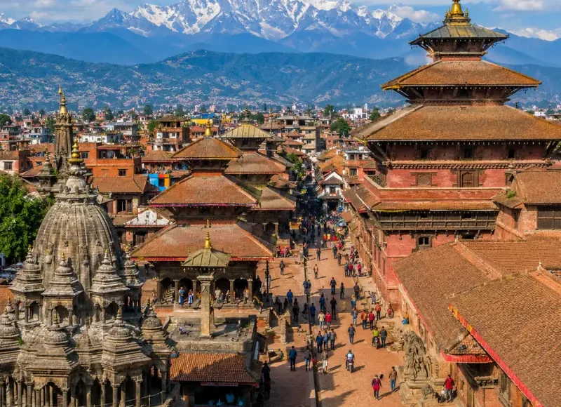 Bright daytime view of Kathmandu with traditional temples, historic square, city rooftops, and Himalayan mountains.
