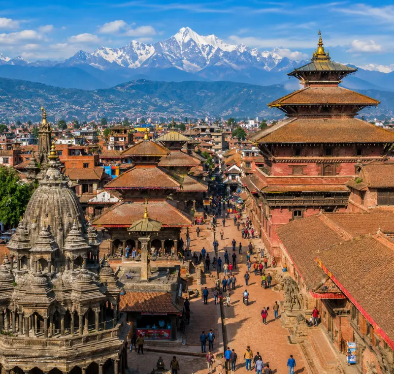 Bright daytime view of Kathmandu with traditional temples, historic square, city rooftops, and Himalayan mountains.