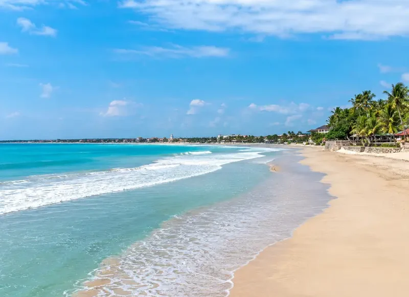 Kuta Beach in Bali with turquoise waves, golden sand, palm trees, and clear blue daytime sky.