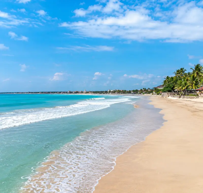 Kuta Beach in Bali with turquoise waves, golden sand, palm trees, and clear blue daytime sky.