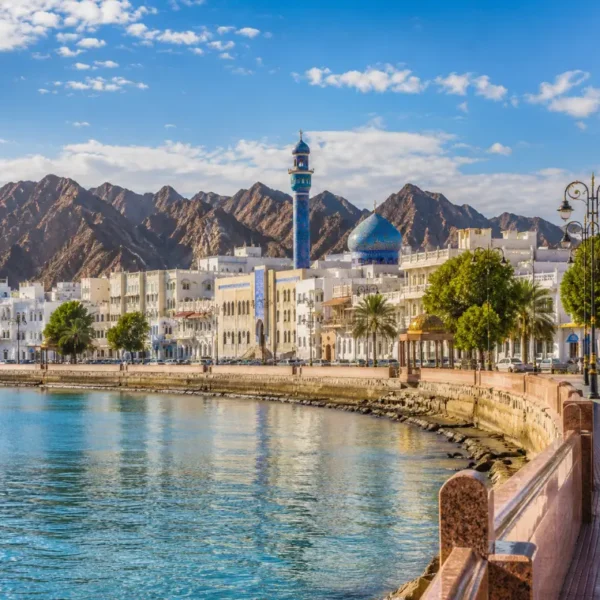 Bright daytime view of Muscat waterfront with calm sea, elegant promenade, traditional architecture, and rugged mountains.