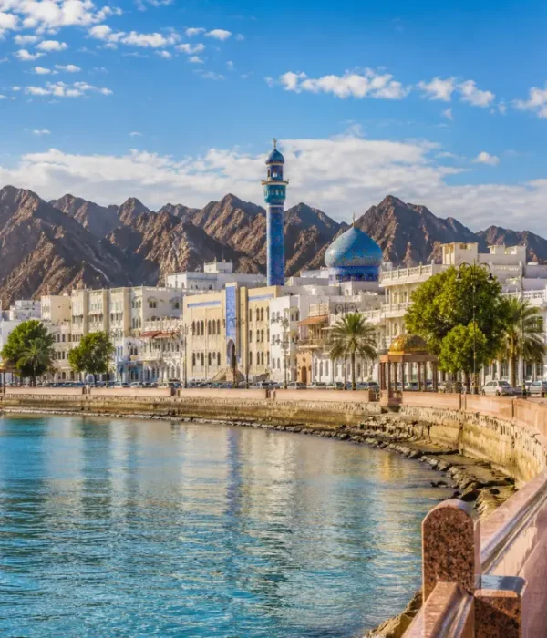 Bright daytime view of Muscat waterfront with calm sea, elegant promenade, traditional architecture, and rugged mountains.
