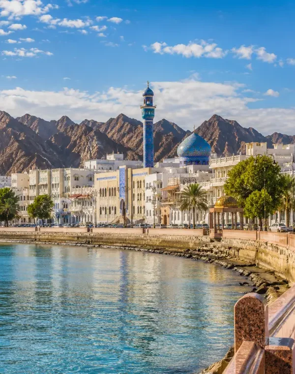 Bright daytime view of Muscat waterfront with calm sea, elegant promenade, traditional architecture, and rugged mountains.