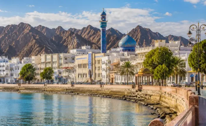 Bright daytime view of Muscat waterfront with calm sea, elegant promenade, traditional architecture, and rugged mountains.