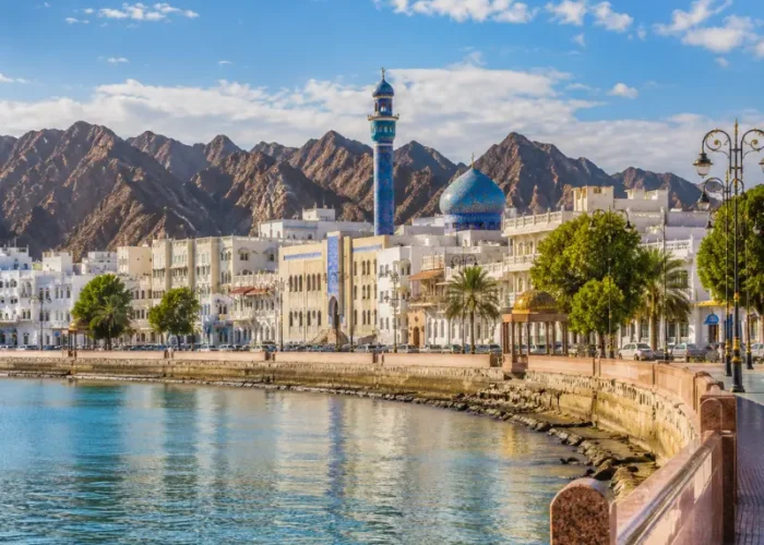 Bright daytime view of Muscat waterfront with calm sea, elegant promenade, traditional architecture, and rugged mountains.