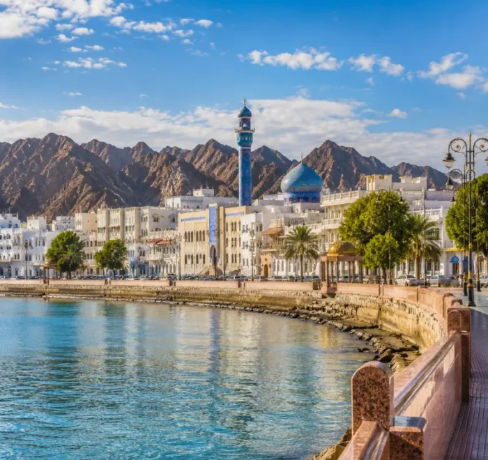 Bright daytime view of Muscat waterfront with calm sea, elegant promenade, traditional architecture, and rugged mountains.
