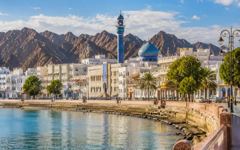 Bright daytime view of Muscat waterfront with calm sea, elegant promenade, traditional architecture, and rugged mountains.