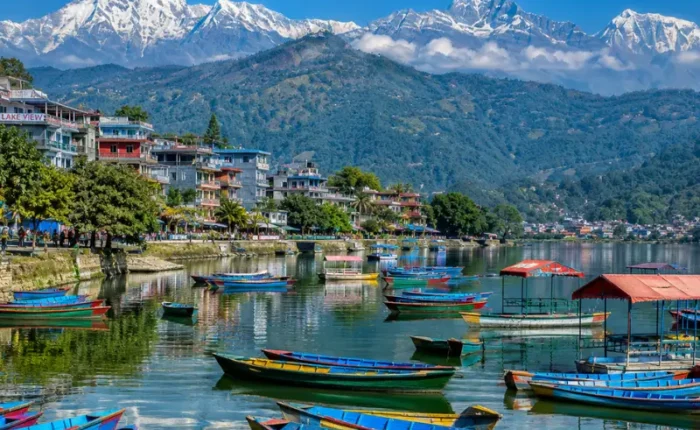 Bright daytime view of Pokhara with Phewa Lake, colorful boats, lakeside town, green hills, and snow-capped mountains.