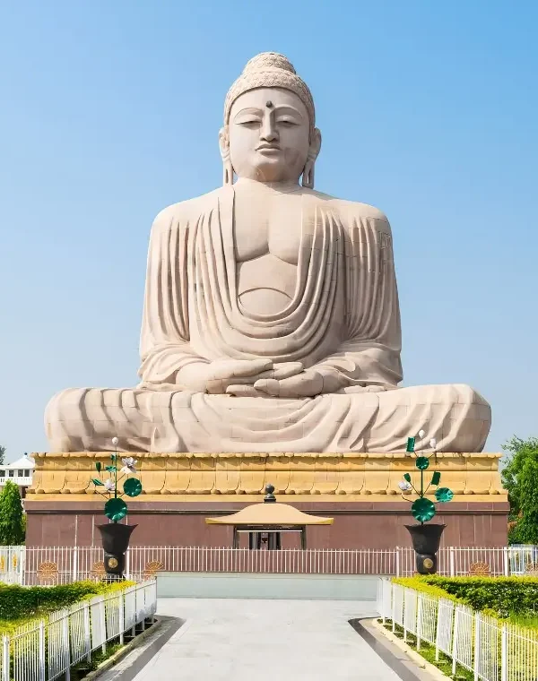 Bodh Gaya travel image featuring a large seated Buddha statue, landscaped gardens, clear blue sky, and peaceful Buddhist pilgrimage surroundings.