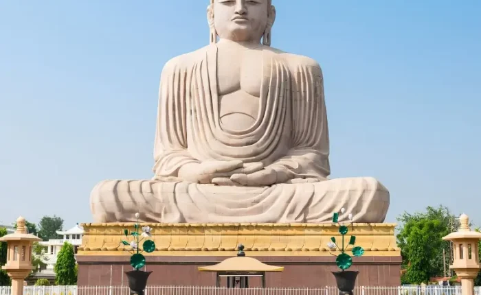 Bodh Gaya travel image featuring a large seated Buddha statue, landscaped gardens, clear blue sky, and peaceful Buddhist pilgrimage surroundings.
