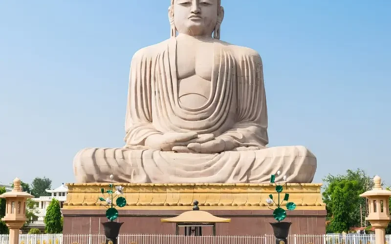 Bodh Gaya travel image featuring a large seated Buddha statue, landscaped gardens, clear blue sky, and peaceful Buddhist pilgrimage surroundings.
