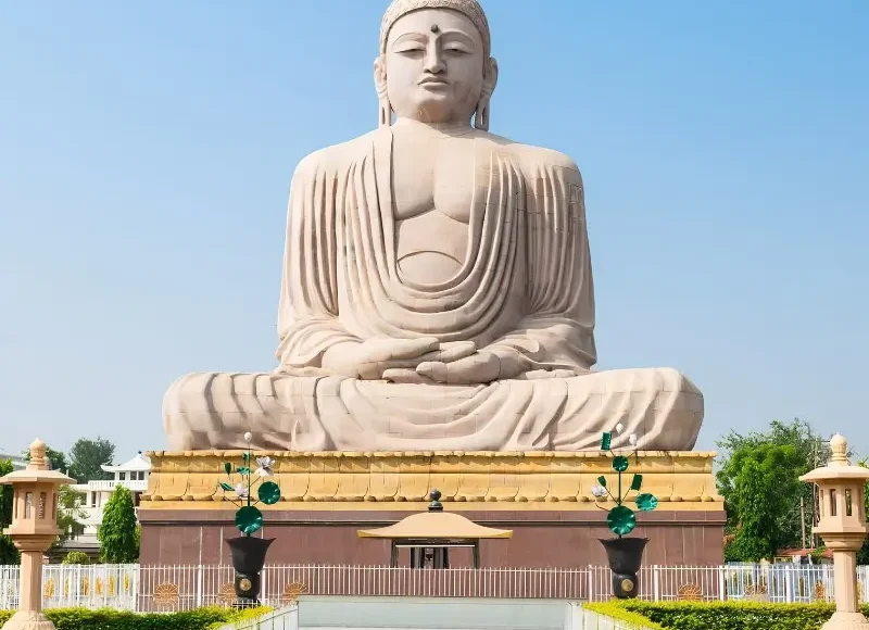 Bodh Gaya travel image featuring a large seated Buddha statue, landscaped gardens, clear blue sky, and peaceful Buddhist pilgrimage surroundings.