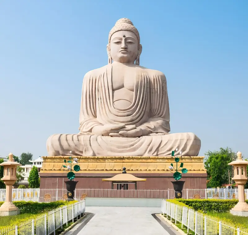 Bodh Gaya travel image featuring a large seated Buddha statue, landscaped gardens, clear blue sky, and peaceful Buddhist pilgrimage surroundings.