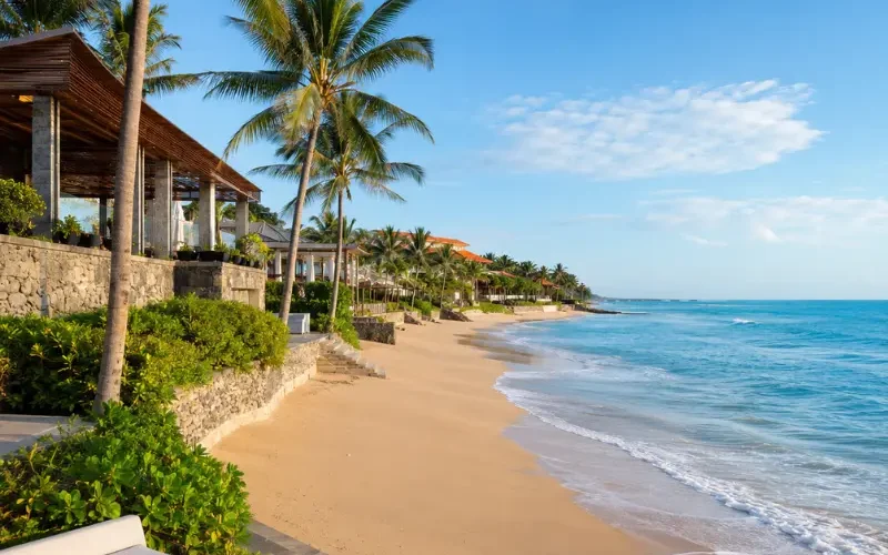 Seminyak, Bali beachfront with golden sand, turquoise sea, swaying palm trees, and luxury seaside villas in clear daylight.