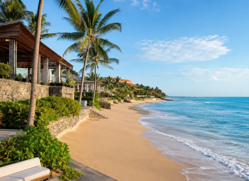 Seminyak, Bali beachfront with golden sand, turquoise sea, swaying palm trees, and luxury seaside villas in clear daylight.
