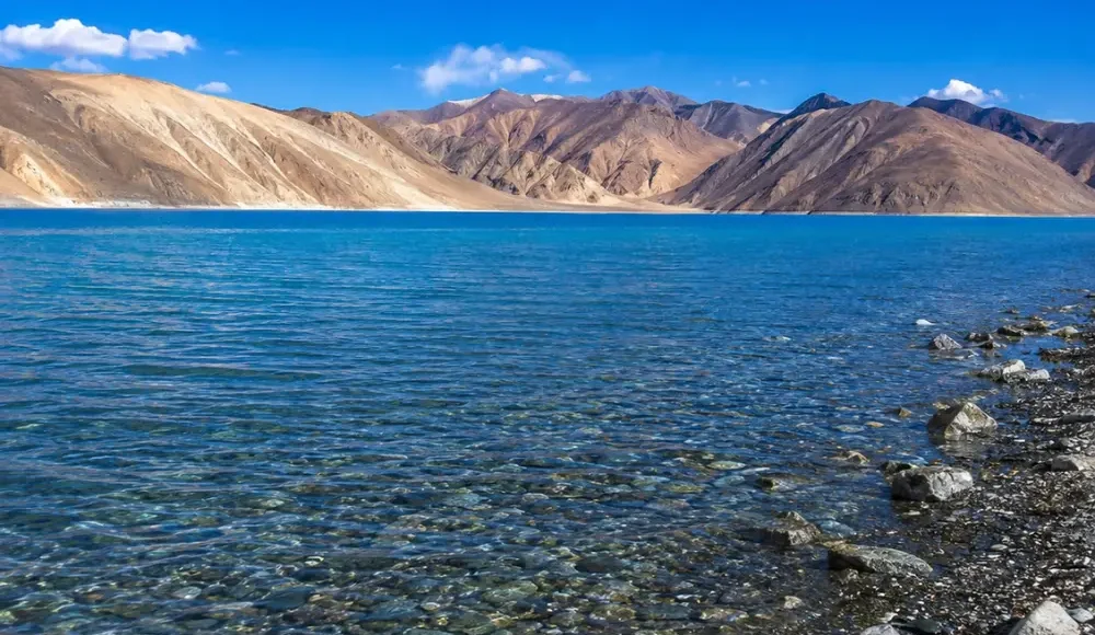 Crystal-clear Pangong Lake in Ladakh with turquoise blue water, rocky shoreline, and barren Himalayan mountains.