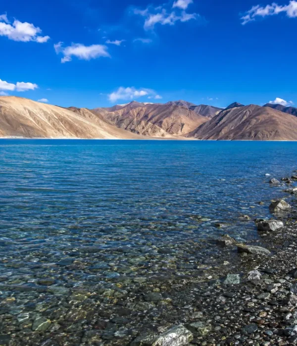 Crystal-clear Pangong Lake in Ladakh with turquoise blue water, rocky shoreline, and barren Himalayan mountains.