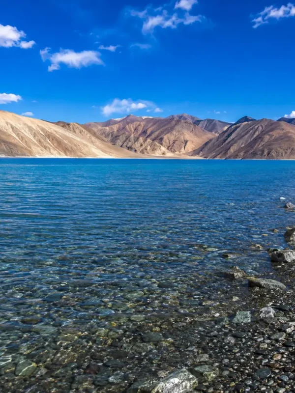 Crystal-clear Pangong Lake in Ladakh with turquoise blue water, rocky shoreline, and barren Himalayan mountains.