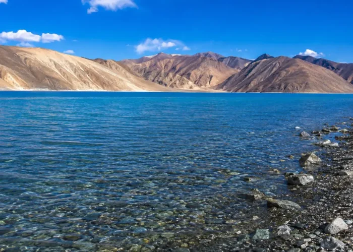 Crystal-clear Pangong Lake in Ladakh with turquoise blue water, rocky shoreline, and barren Himalayan mountains.