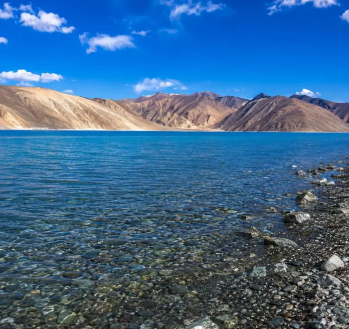 Crystal-clear Pangong Lake in Ladakh with turquoise blue water, rocky shoreline, and barren Himalayan mountains.