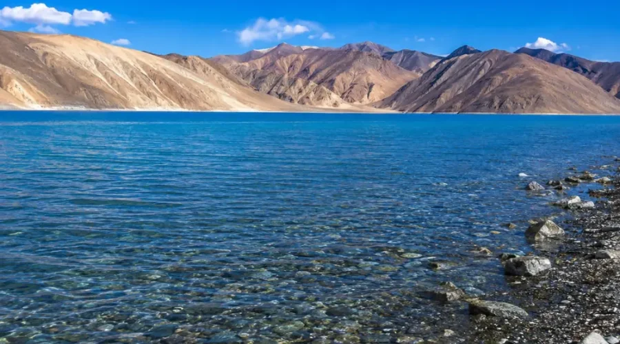 Crystal-clear Pangong Lake in Ladakh with turquoise blue water, rocky shoreline, and barren Himalayan mountains.