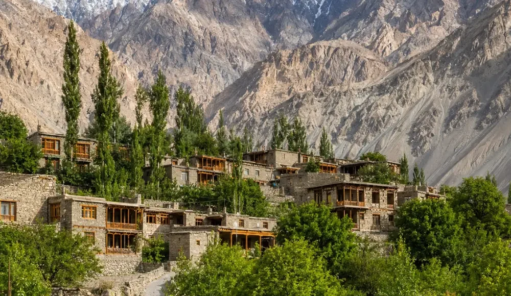 Scenic Turtuk village in Ladakh with traditional stone houses, wooden bridge, green trees, and rugged mountain peaks.