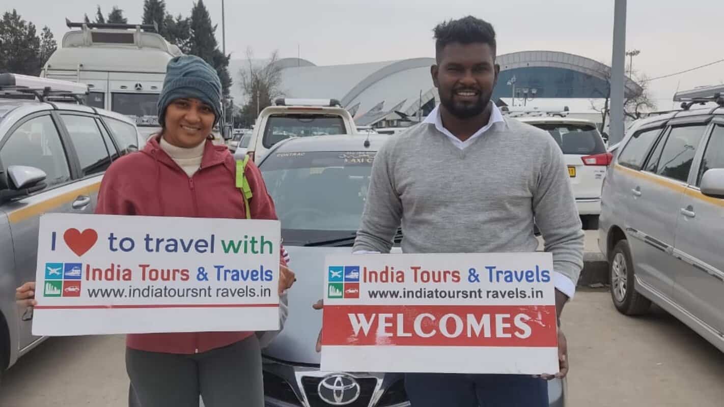 Travelers welcoming guests with India Tours & Travels signage at a busy parking lot in India.