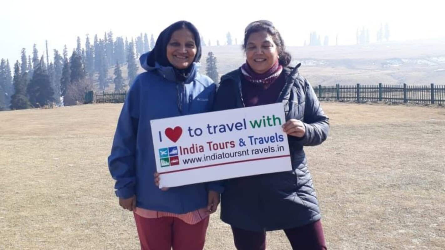 Beautiful women holding a sign promoting India Tours & Travels in scenic outdoor location, showcasing travel experiences in India for adventurers and cultural explorers.