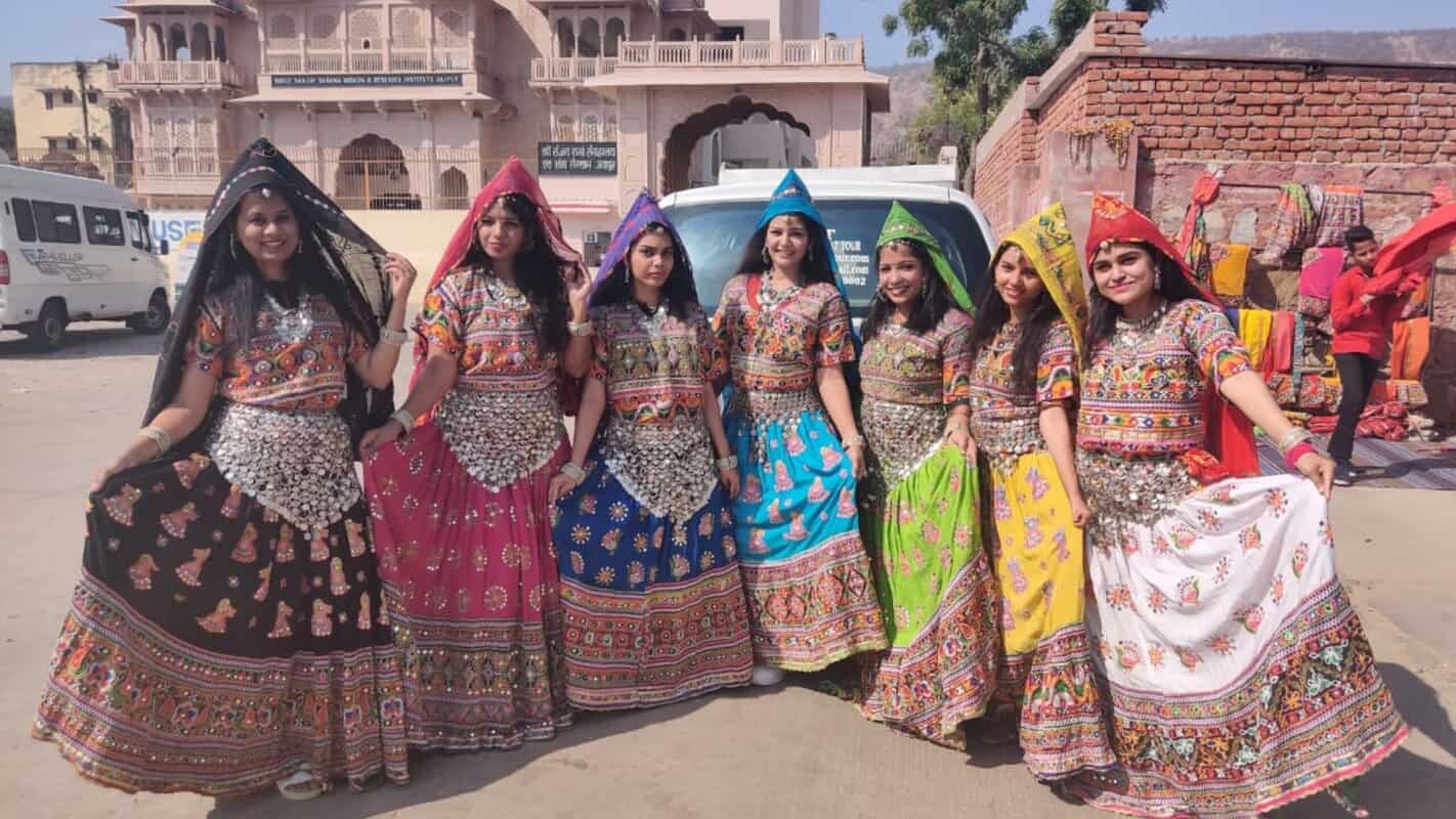 Vibrant group of women dressed in traditional Rajasthani attire during a cultural event in India, showcasing colorful lehengas and turbans, with historic architecture in the background.